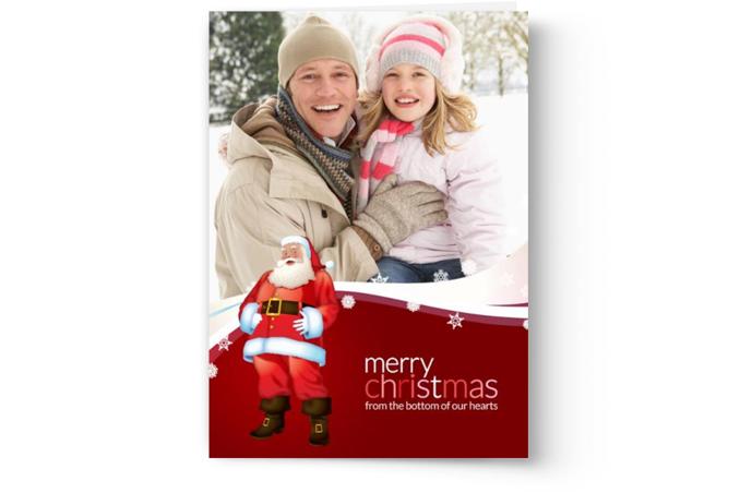 A holiday greeting card from PhotoBook Press featuring a man and a child smiling in a winter setting with a Santa Claus figurine and the message "Merry Christmas from the bottom of our hearts.