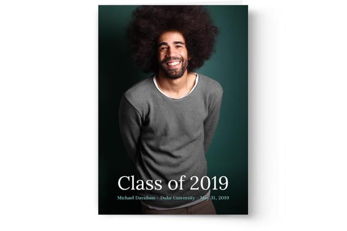 A man with afro hair is smiling in front of a green background, holding Photo Book Press's personalized graduation announcement cards and invitations.