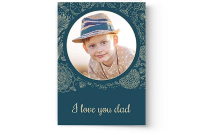 A personalized Photo Book Press Father’s Day card featuring a photo of a young child wearing a hat with the text “I love you dad” on a dark blue background with floral patterns.